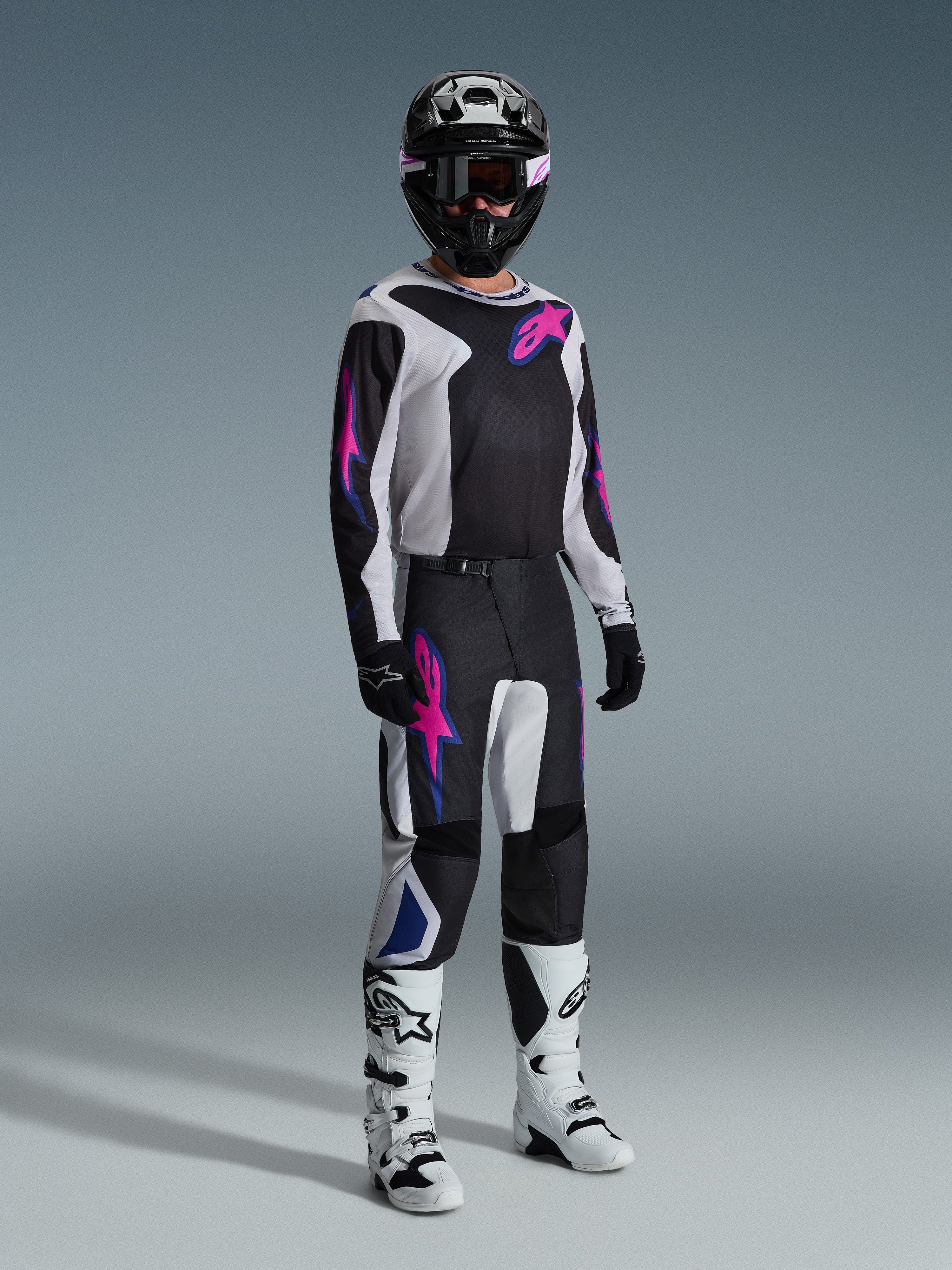 Piloto de motocross con equipamiento completo de carreras, Jersey Fluid Grid 2026 en negro con detalles en gris claro y púrpura, pantalones Fluid Grid a juego, casco negro con gafas coordinadas, botas blancas y negras, y guantes negros, en un estudio profesional para carreras off-road.