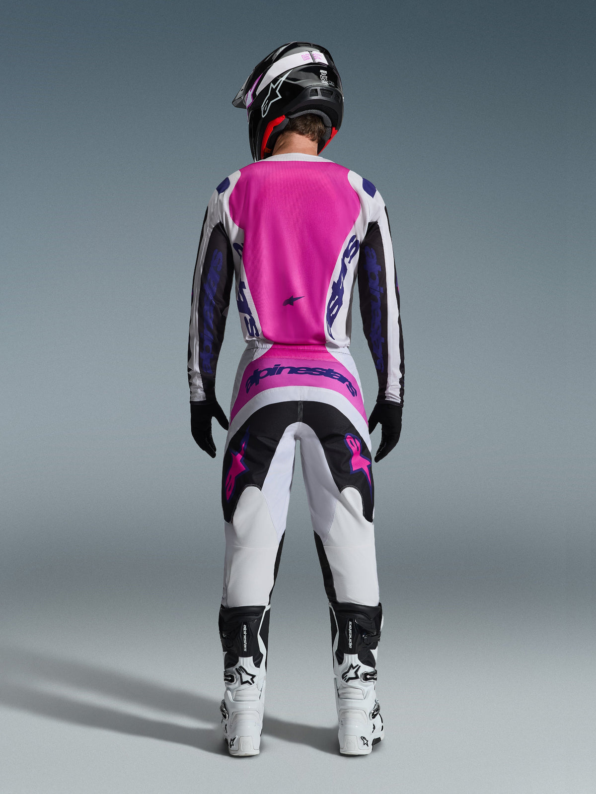 Alpinestars 2026 Fluid Grid Gear Set, piloto de motocross desde atrás con jersey rosa brillante y blanco de mangas negras, pantalones blancos a juego con paneles negros y rosas, logotipo de Alpinestars en púrpura, botas de carreras blancas y negras, y casco coordinado, diseñado para el rendimiento off-road