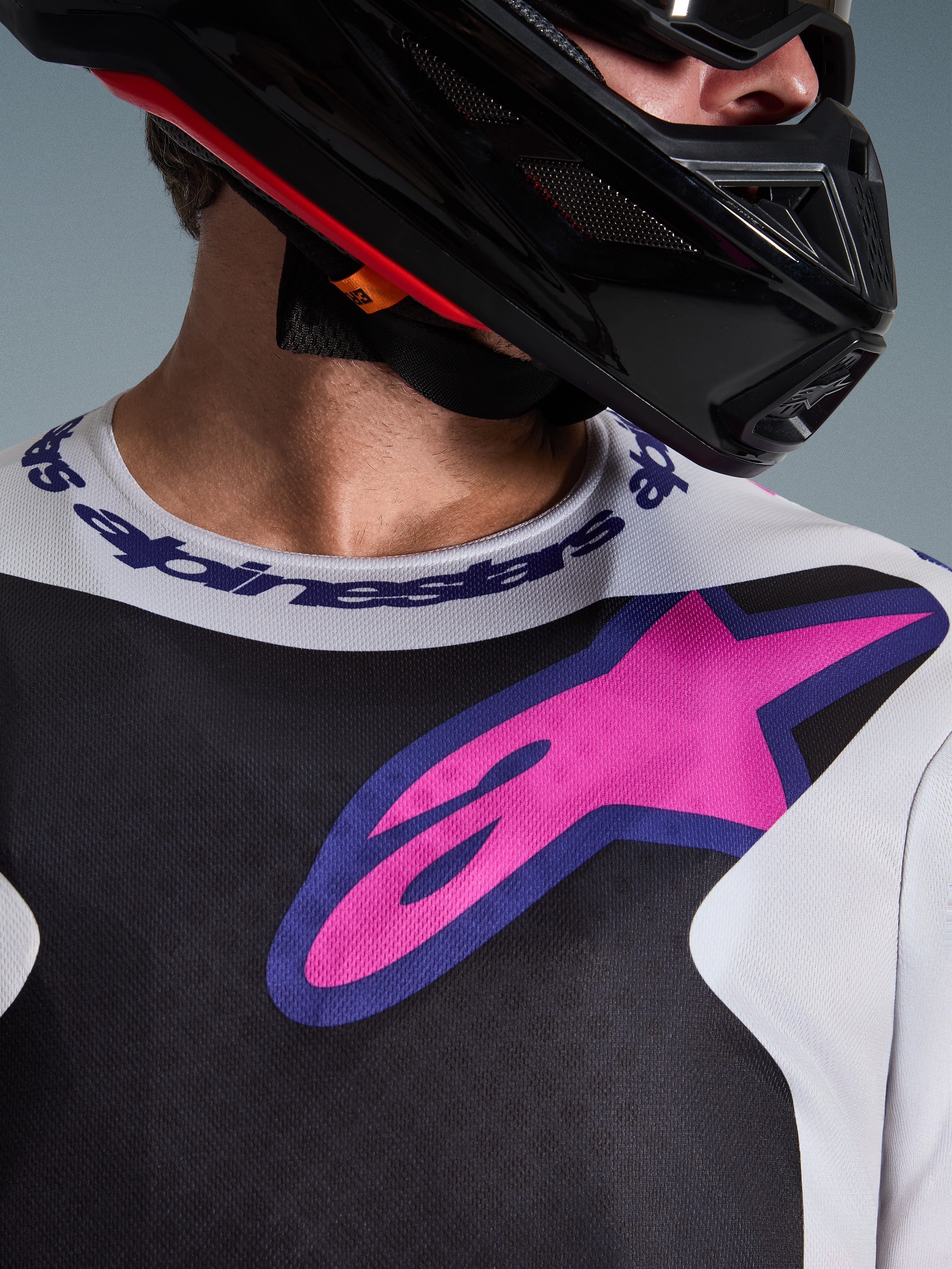 Alpinestars 2026 Fluid Grid Jersey, jersey de motocross, negro, gris claro y morado, primer plano del área del pecho y el cuello con un gran logotipo de estrella rosa, cuello deportivo de marca, usado con casco negro, equipamiento off-road de alto rendimiento