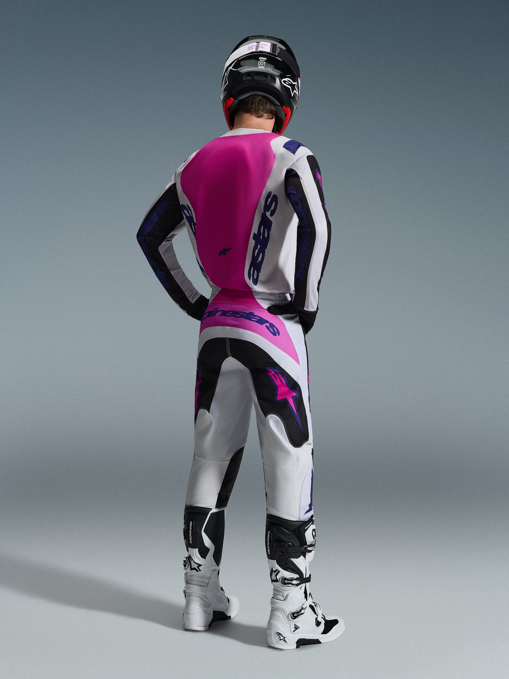 Piloto de motocross desde atrás vistiendo el Jersey Alpinestars Fluid Grid 2026, jersey blanco con un gran panel trasero en rosa fucsia y logotipos morados, pantalones blancos y negros a juego con detalles rosas, botas blancas y casco negro, equipamiento coordinado para carreras off-road