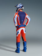 Motocross rider from behind wearing Alpinestars 2026 Fluid Grid Gear Set, Orange and UCLA Blue jersey with white sleeves, matching orange and white pants, blue motocross boots, and blue and white helmet, coordinated performance outfit for off-road racing