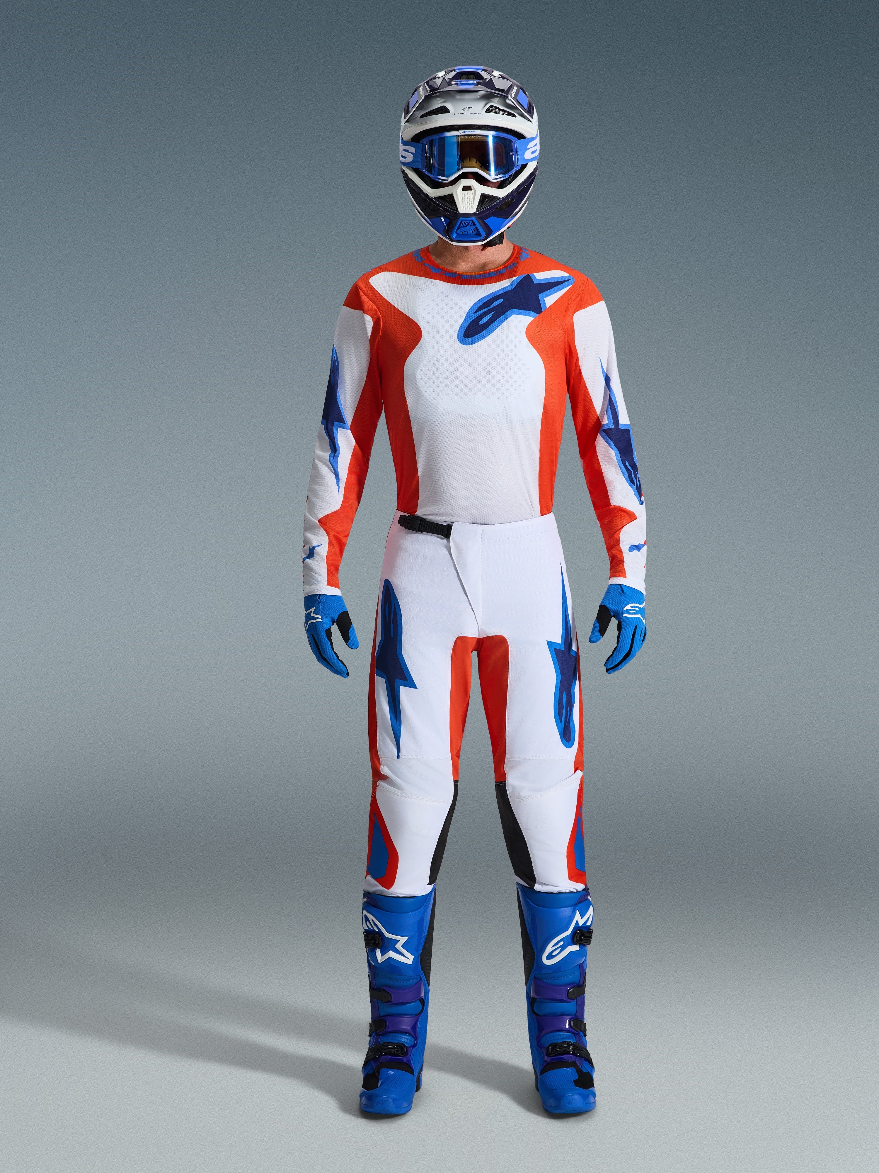 Alpinestars 2026 Fluid Grid Gear Set, piloto de motocross con equipamiento completo, jersey y pantalones blancos con acentos en naranja y azul UCLA, casco azul y blanco a juego con gafas azules, guantes azules y botas de motocross azules, atuendo coordinado para carreras todoterreno