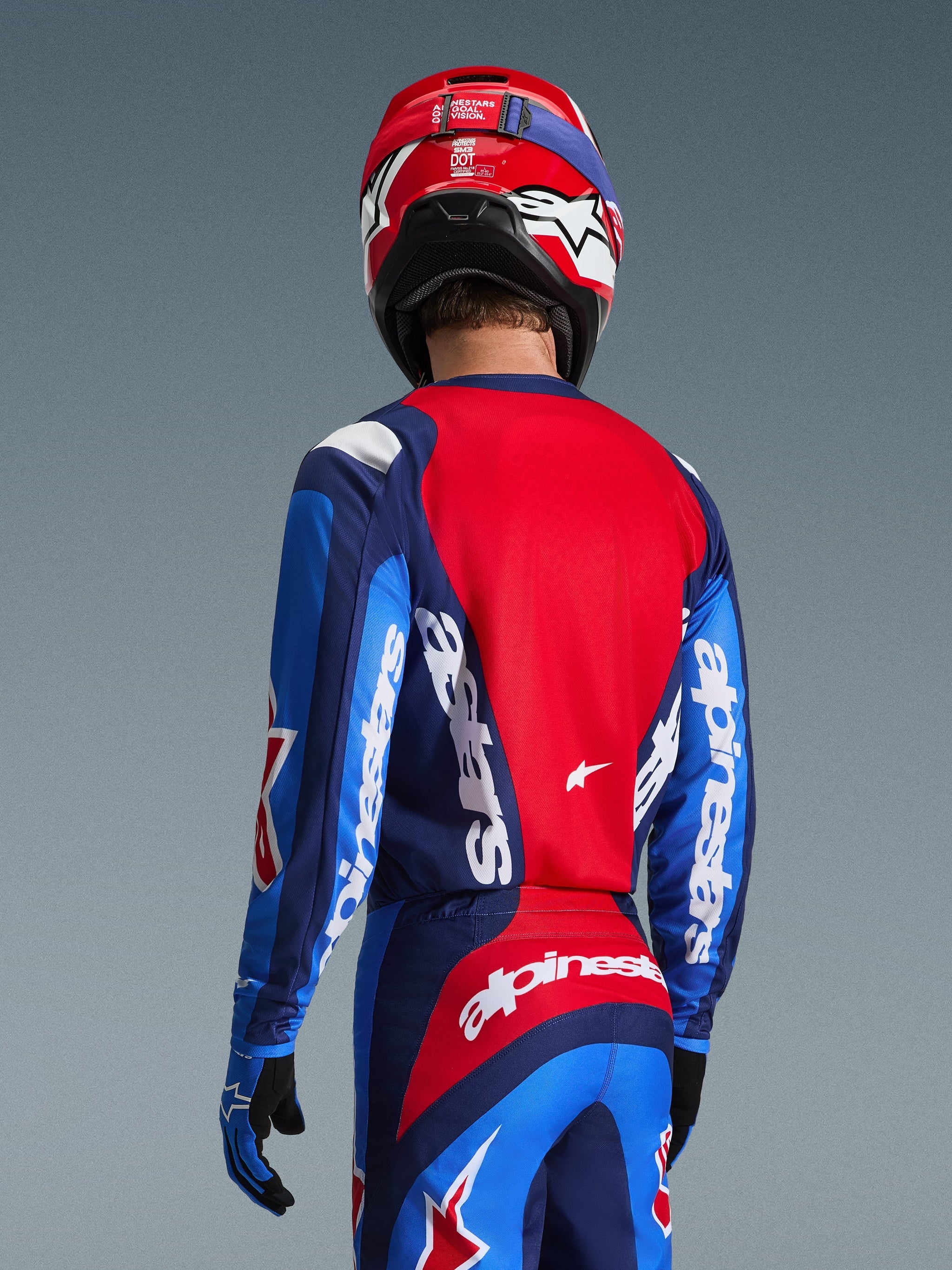 Vista trasera de un piloto de motocross con el Alpinestars 2026 Fluid Grid Jersey en azul y rojo, con logotipos blancos llamativos en las mangas y la espalda, combinado con pantalones de motocross a juego y un casco rojo para un look completo de carreras off-road