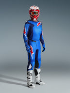 Piloto de motocross con el Jersey Alpinestars Fluid Grid 2026 en azul y rojo, combinado con pantalones de motocross a juego, botas blancas y negras y un casco rojo y blanco, conjunto de equipamiento profesional para rendimiento off-road.