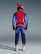 Vista posterior de un piloto de motocross con el Jersey Fluid Grid 2026 en azul y rojo, con un gran panel trasero rojo y mangas azules con la marca Alpinestars en blanco, combinado con pantalones a juego, botas blancas y negras, y un casco rojo para un equipo completo de motocross.
