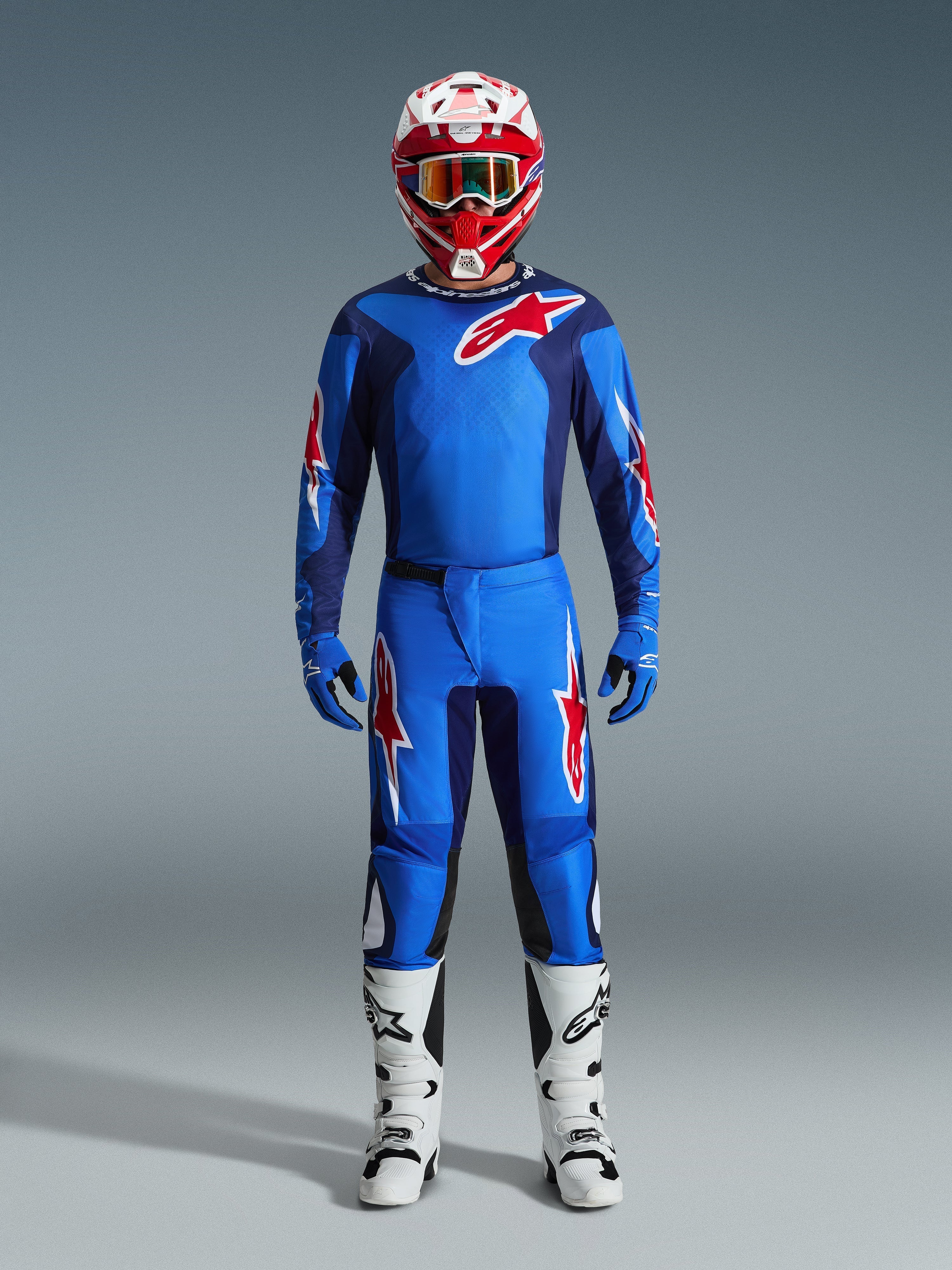 Piloto de motocross con el Jersey Alpinestars Fluid Grid 2026 en azul y rojo, pantalones azules a juego con logotipos de estrellas blancas y rojas, casco rojo y blanco con gafas de espejo, guantes azules y botas de carreras blancas y negras, conjunto completo de equipamiento para todoterreno.
