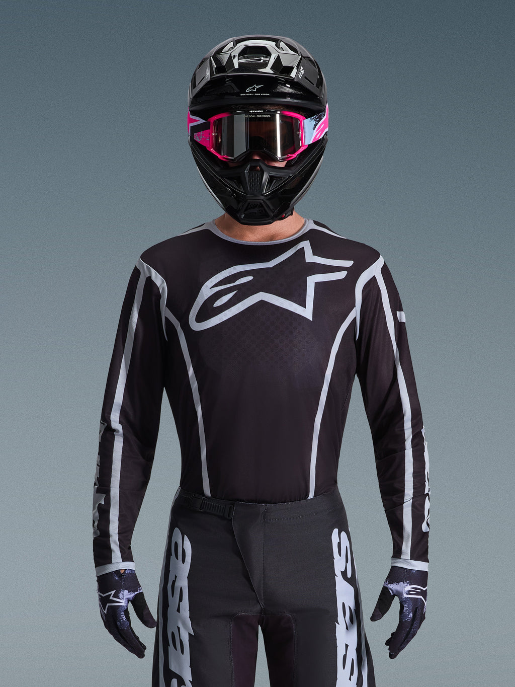 Alpinestars Jersey Fluid Apex 2026, jersey de motocross, negro y gris, con un gran logotipo Astars blanco en el pecho y detalles gráficos grises en las mangas, usado por un piloto con gafas rosas y casco negro, equipamiento de carreras off-road coordinado