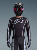 Alpinestars Jersey Fluid Apex 2026, jersey de motocross, negro y gris, con un gran logotipo Astars blanco en el pecho y detalles gráficos grises en las mangas, usado por un piloto con gafas rosas y casco negro, equipamiento de carreras off-road coordinado