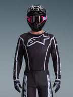 Alpinestars Jersey Fluid Apex 2026, jersey de motocross, negro y gris, con un gran logotipo Astars blanco en el pecho y detalles gráficos grises en las mangas, usado por un piloto con gafas rosas y casco negro, equipamiento de carreras off-road coordinado