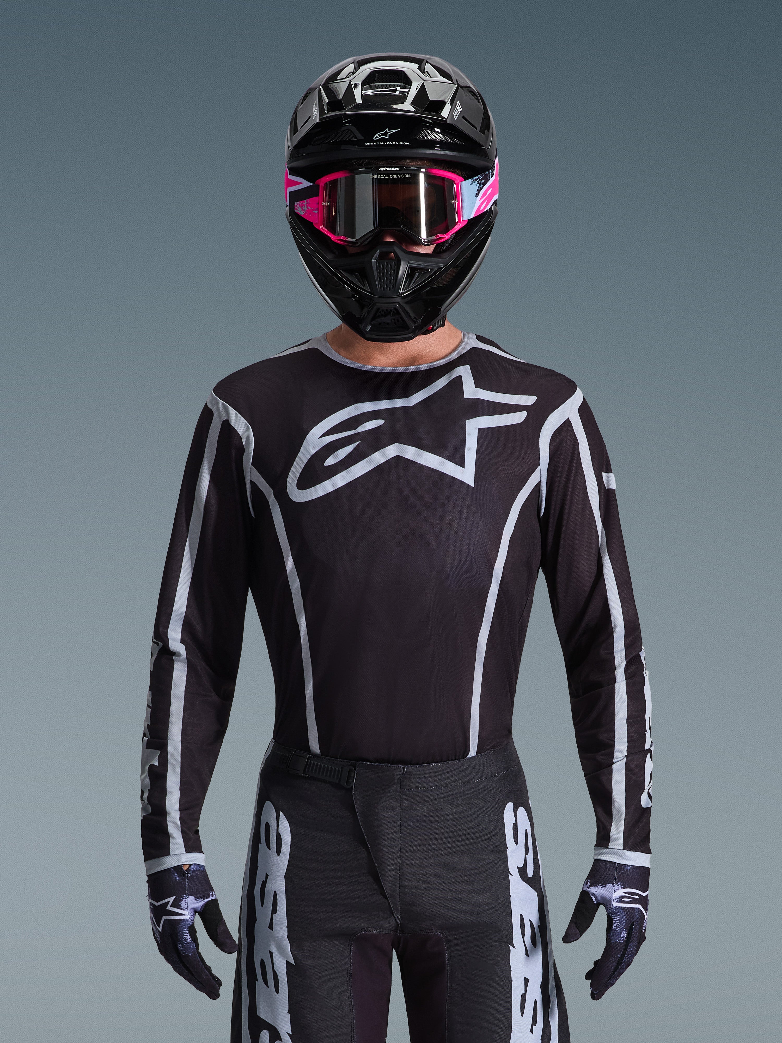 Alpinestars Jersey Fluid Apex 2026, jersey de motocross, negro y gris, con un gran logotipo Astars blanco en el pecho y detalles gráficos grises en las mangas, usado por un piloto con gafas rosas y casco negro, equipamiento de carreras off-road coordinado