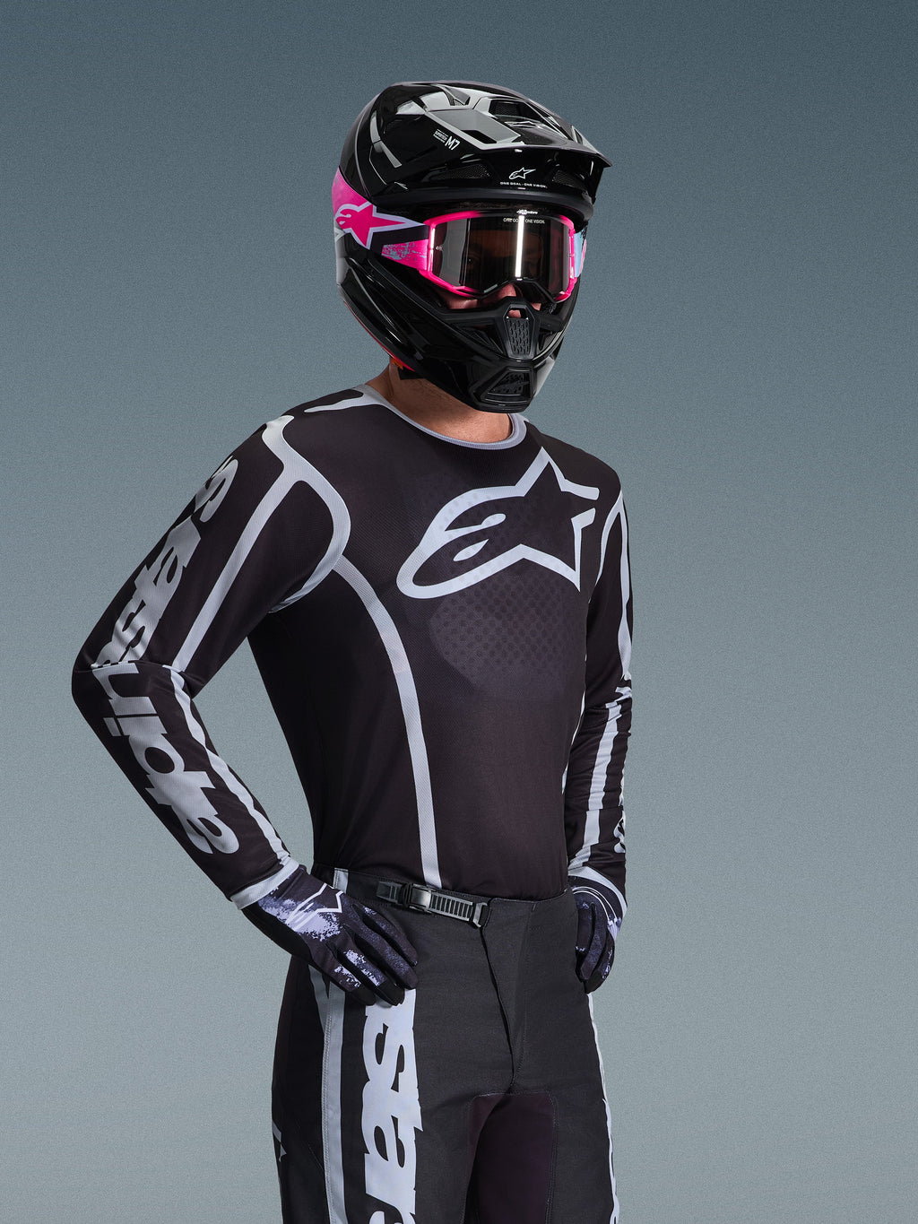 Piloto de motocross vistiendo el Jersey Alpinestars Fluid Apex 2026, negro y gris con un logotipo blanco grande en el pecho y gráficos geométricos en las mangas, combinado con pantalones negros, guantes negros y un casco negro con acentos y gafas rosas.
