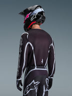 Piloto de motocross visto desde atrás vistiendo el jersey Alpinestars Fluid Apex 2026 en color negro y gris, pantalones de motocross a juego con detalles blancos, casco negro con logotipos en rosa, conjunto coordinado para carreras off-road.