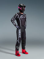 Piloto de motocross con el Jersey Fluid Apex 2026 en color negro y gris, combinado con pantalones a juego, casco negro con gafas rosas y botas de color rojo vibrante, con la llamativa marca Alpinestars en blanco en el pecho y las mangas, conjunto profesional coordinado de carreras off-road.