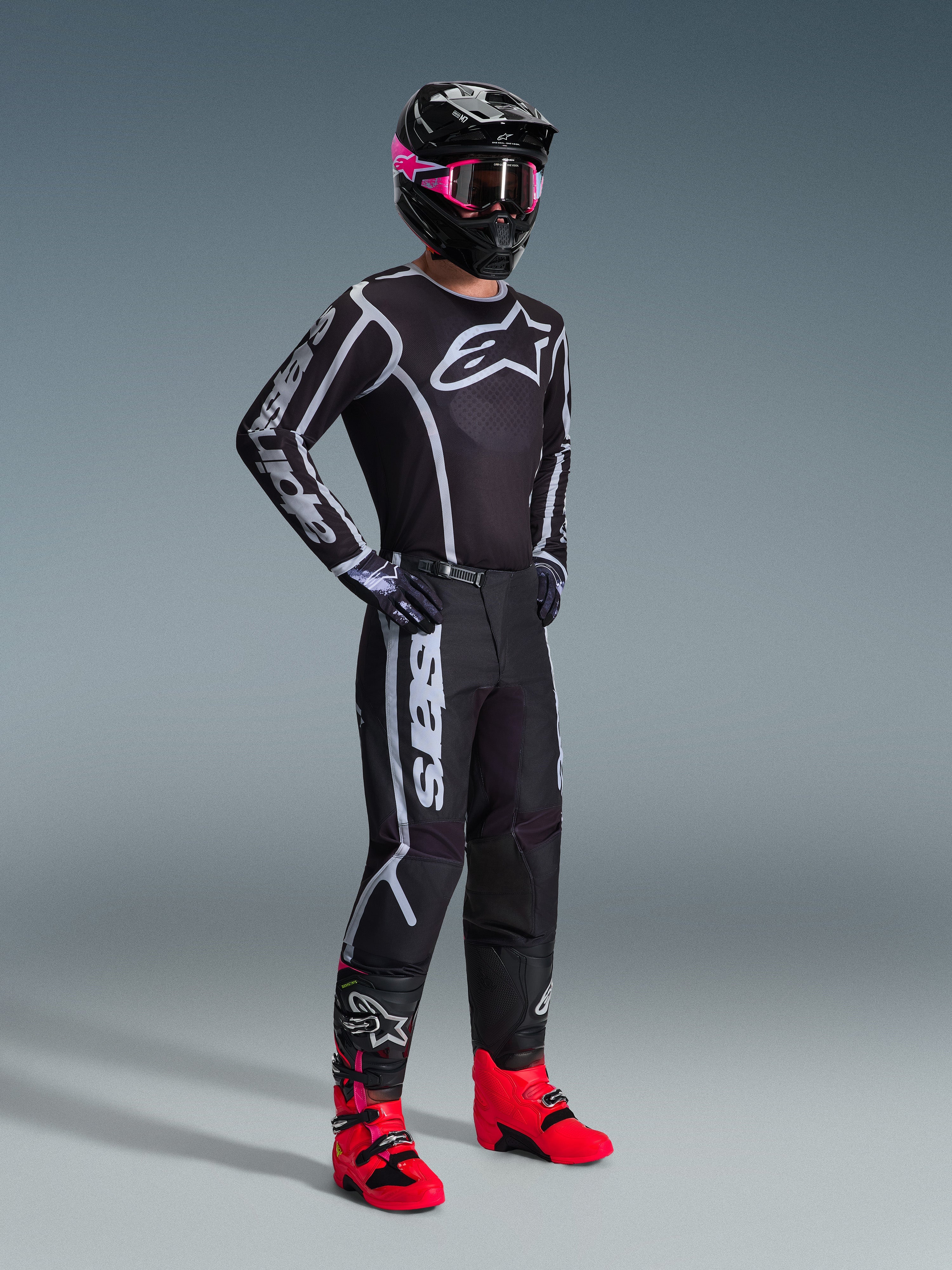 Piloto de motocross con el Jersey Fluid Apex 2026 en color negro y gris, combinado con pantalones a juego, casco negro con gafas rosas y botas de color rojo vibrante, con la llamativa marca Alpinestars en blanco en el pecho y las mangas, conjunto profesional coordinado de carreras off-road.