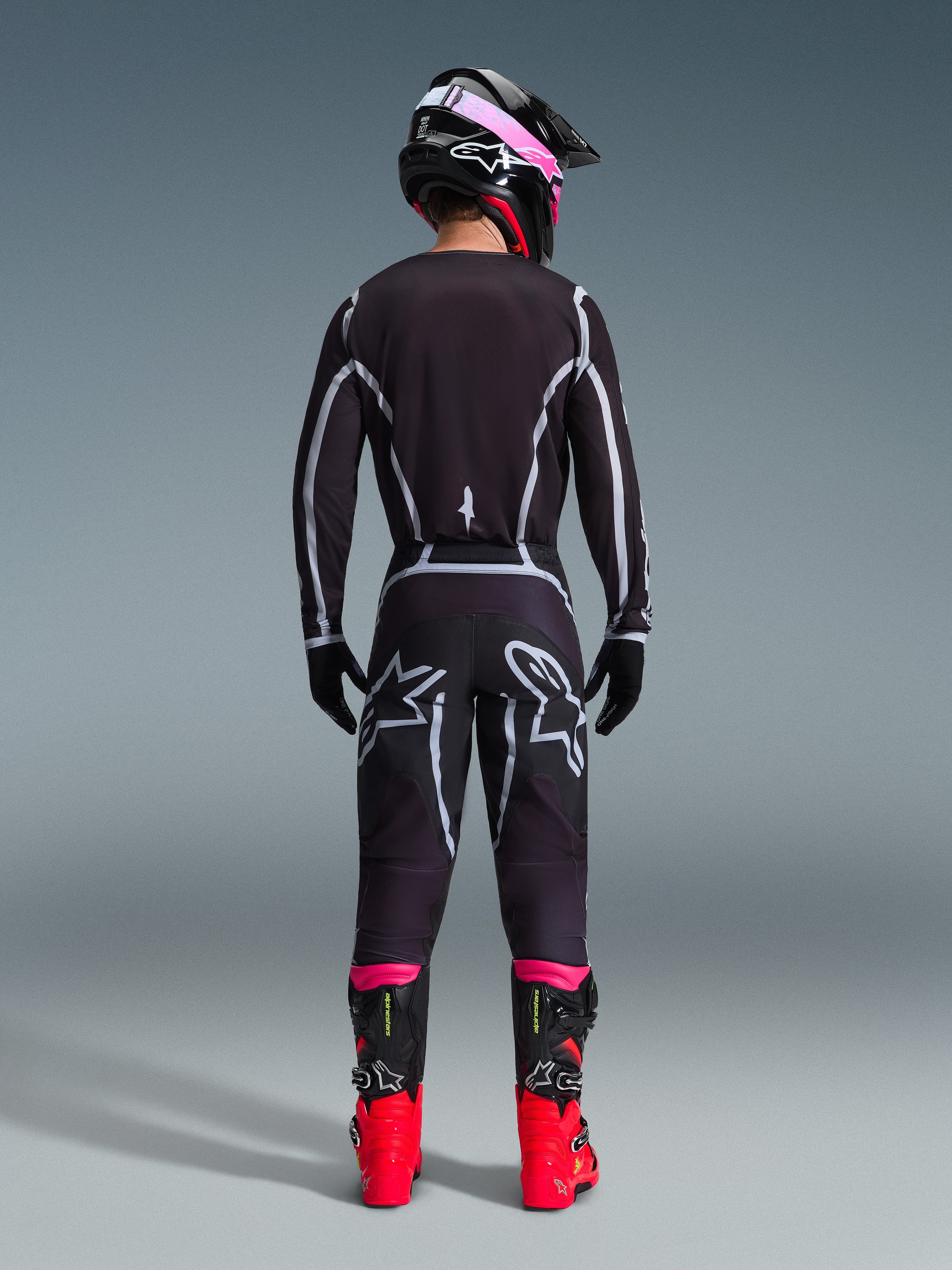 Piloto de motocross visto desde atrás con el conjunto Alpinestars 2026 Fluid Apex, jersey y pantalones negros con líneas geométricas grises y logotipos de estrella, casco negro con detalles rosa, botas rojo brillante y negro, equipo de carreras de motocross coordinado