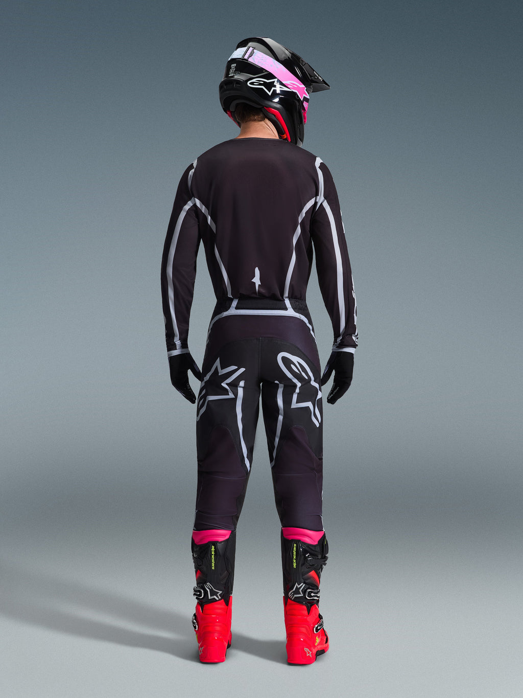 Piloto de motocross desde atrás con el Jersey Alpinestars 2026 Fluid Apex y pantalones a juego en negro/gris, con líneas de diseño geométricas blancas y logotipos de estrella en el asiento, complementado con un casco negro y rosa y botas de competición rojas y negras