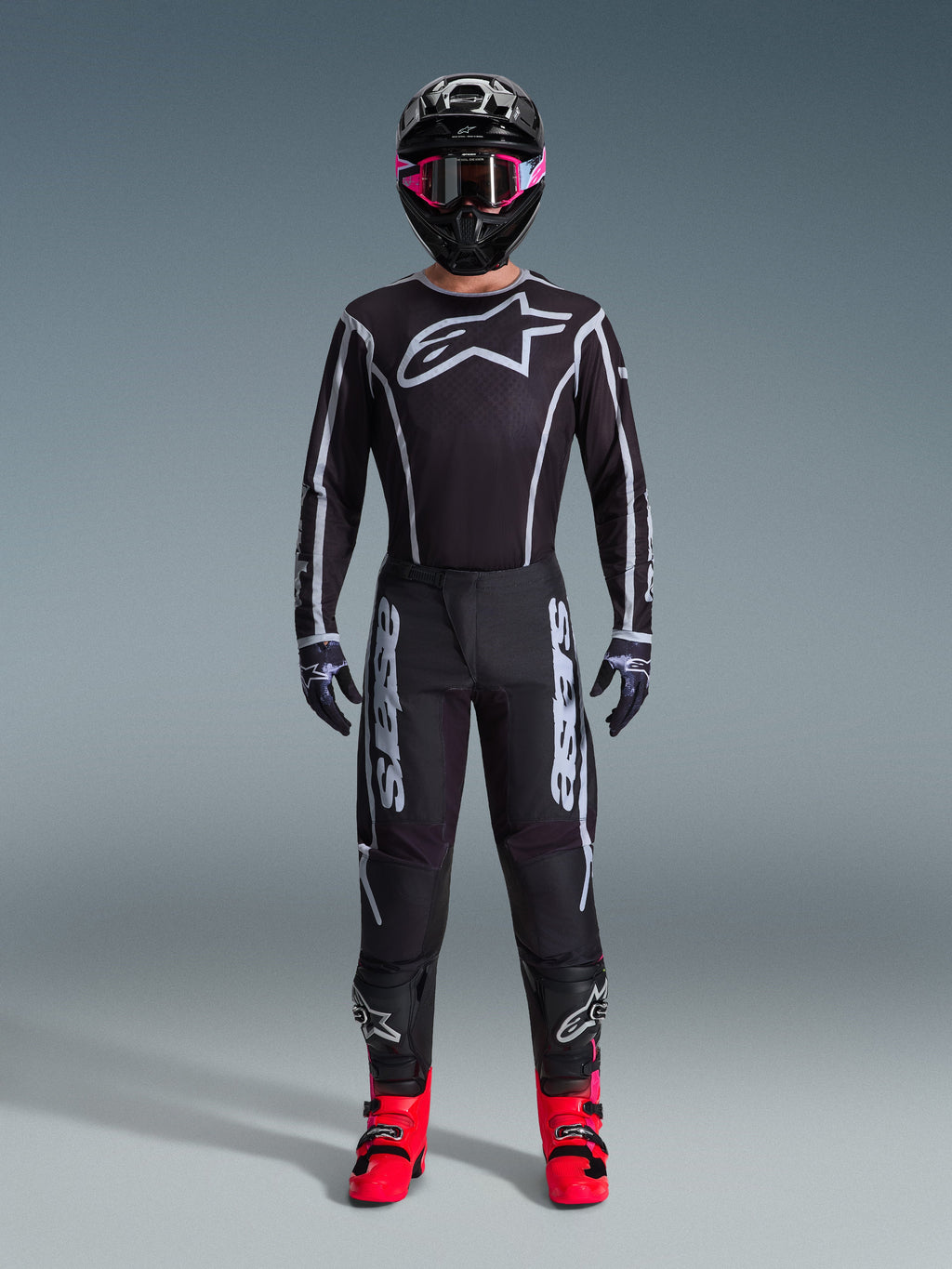 Piloto de motocross con el Jersey Alpinestars Fluid Apex 2026, negro y gris, gran logo blanco de Astars en el pecho, líneas geométricas blancas, pantalones negros a juego, casco negro con gafas rosas y botas rosa brillante, equipamiento completo de carreras de motocross