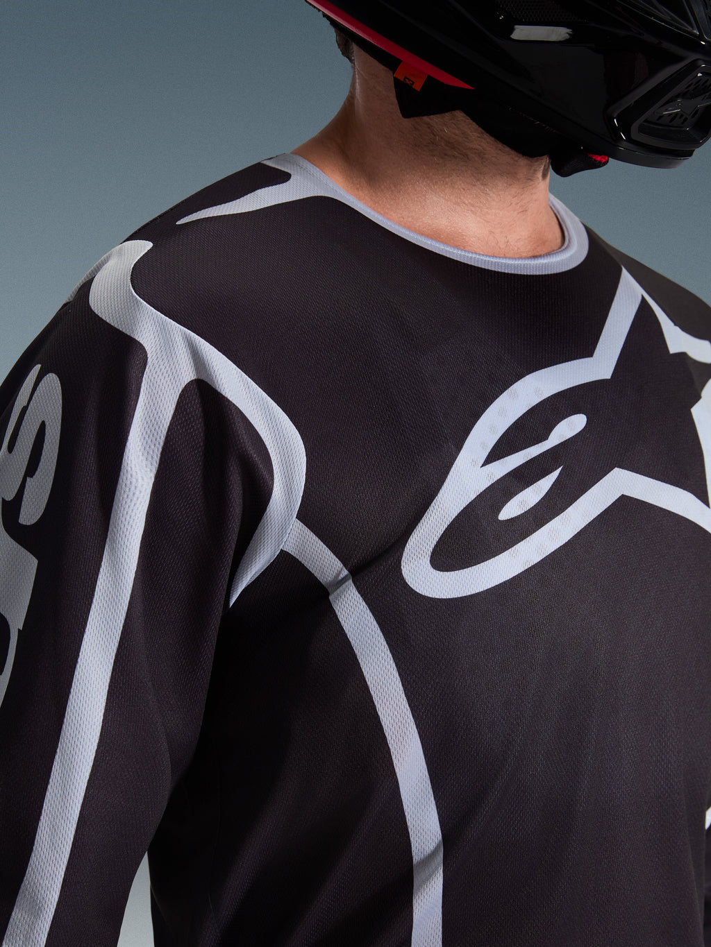 Primer plano de un piloto con el Jersey Alpinestars Fluid Apex 2026 en color negro y gris, que muestra el gran logotipo blanco de Astars en el pecho y la construcción de tejido de malla técnica, combinado con un casco de motocross negro.