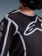Primer plano de un piloto con el Jersey Alpinestars Fluid Apex 2026 en color negro y gris, que muestra el gran logotipo blanco de Astars en el pecho y la construcción de tejido de malla técnica, combinado con un casco de motocross negro.