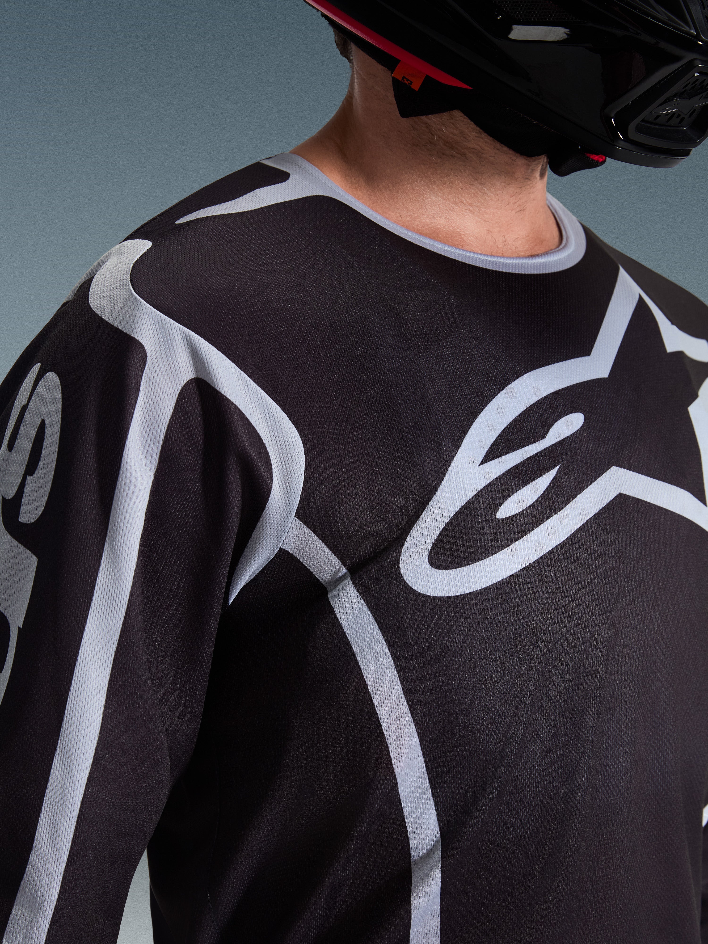 Primer plano de un piloto con el Jersey Alpinestars Fluid Apex 2026 en color negro y gris, que muestra el gran logotipo blanco de Astars en el pecho y la construcción de tejido de malla técnica, combinado con un casco de motocross negro.