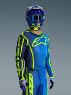 Piloto de motocross con el jersey Alpinestars 2026 Fluid Apex en azul y amarillo flúor, pantalones a juego en azul y amarillo neón, casco púrpura y amarillo con gafas azules y guantes coordinados, equipo completo de carreras todoterreno.