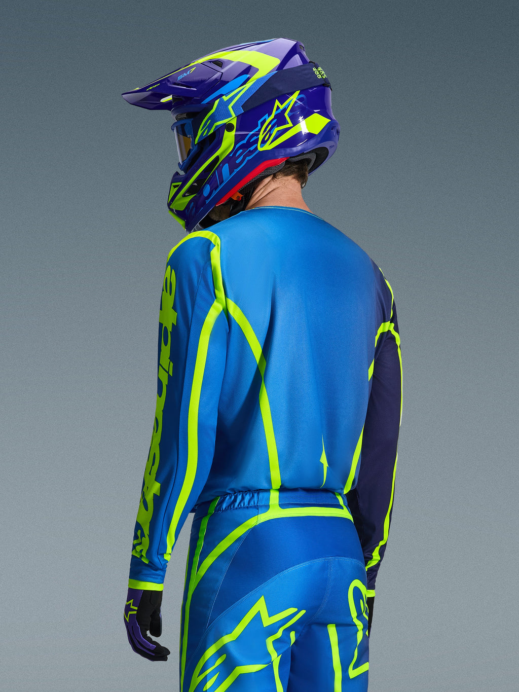 Alpinestars 2026 Fluid Apex Jersey, Azul Amarillo Flúor, vista trasera de un piloto con pantalones de motocross y casco a juego, con gráficos geométricos en amarillo fluorescente y un ajuste atlético relajado, equipamiento coordinado para carreras off-road