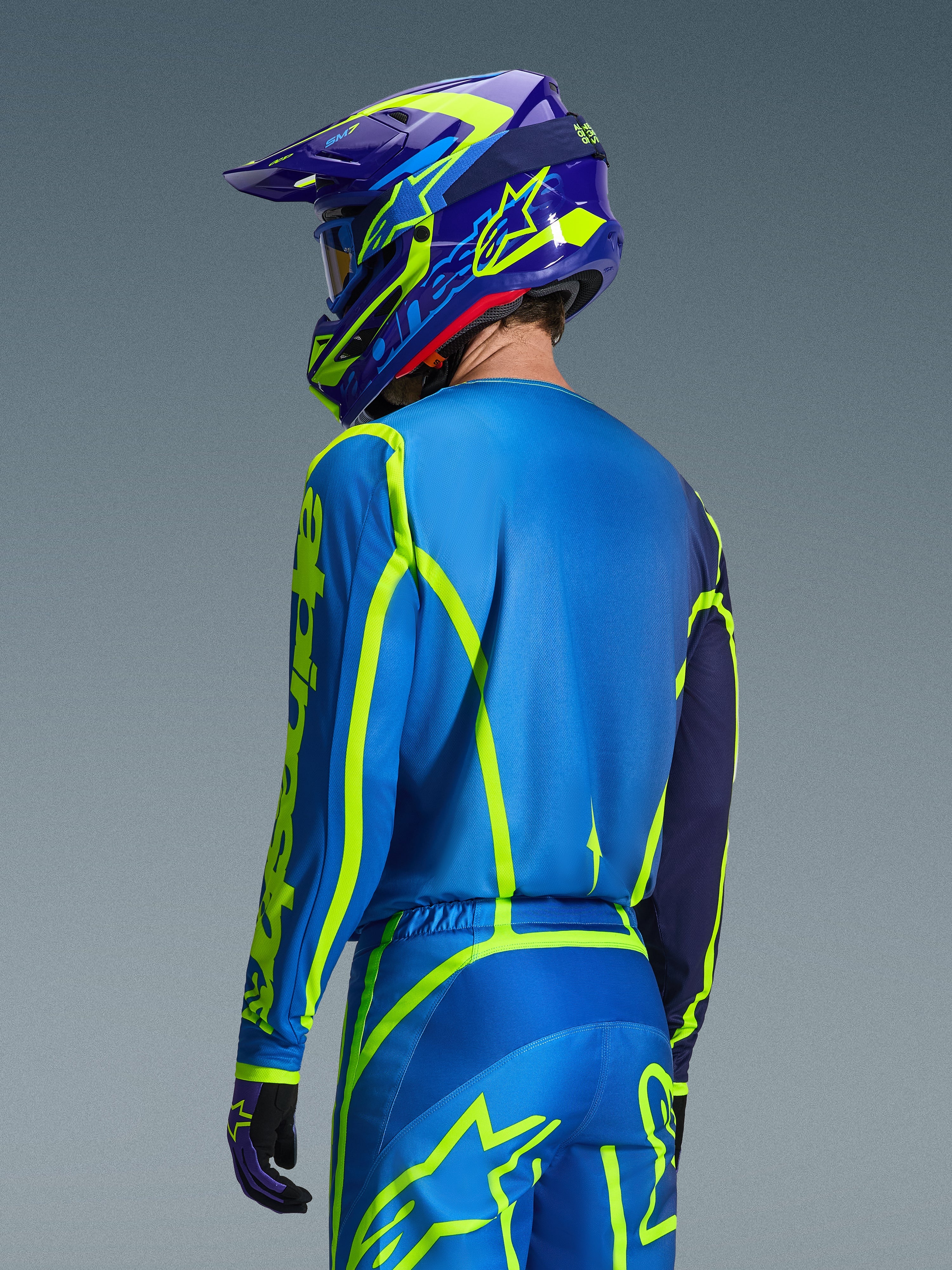 Alpinestars 2026 Fluid Apex Jersey, Azul Amarillo Flúor, vista trasera de un piloto con pantalones de motocross y casco a juego, con gráficos geométricos en amarillo fluorescente y un ajuste atlético relajado, equipamiento coordinado para carreras off-road