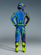 Piloto de motocross visto desde atrás con equipamiento completo de competición, que incluye el Jersey Alpinestars 2026 Fluid Apex y pantalones a juego en Blue Yellow Fluo, con detalles neón vibrantes y logotipos llamativos, combinado con botas amarillo brillante y casco a juego.