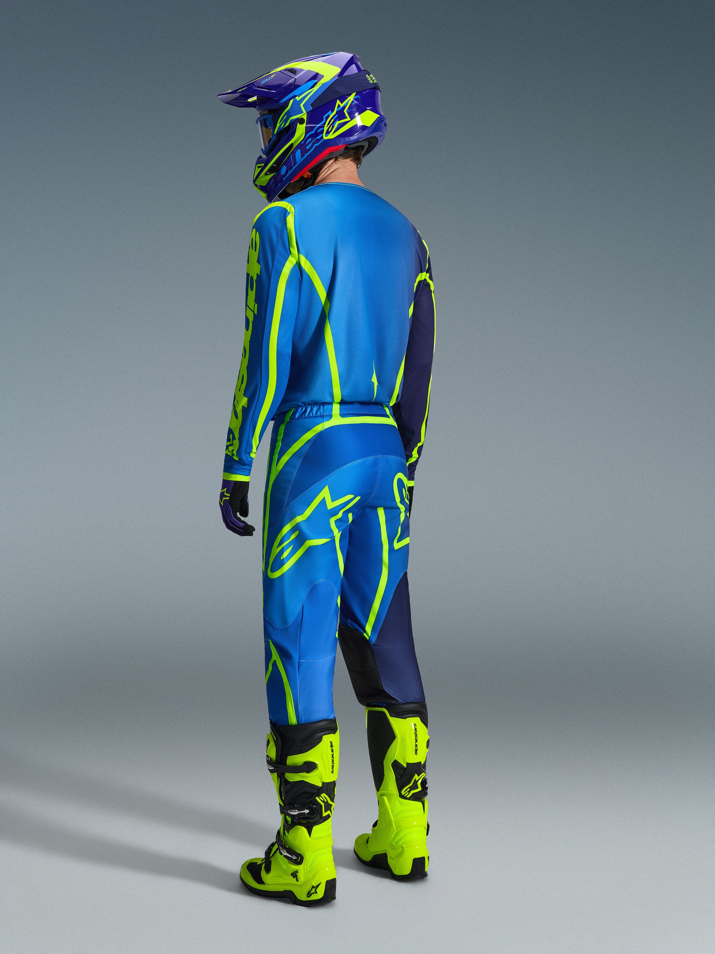 Piloto de motocross visto desde atrás con el jersey Alpinestars 2026 Fluid Apex en color Blue Yellow Fluo, pantalones a juego en azul y amarillo fluorescente, botas amarillo brillante y casco coordinado; equipamiento profesional para carreras off-road