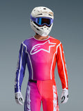 Piloto de motocross con el Jersey Fluid Apex 2026, diseño con degradado vibrante en púrpura, rosa y naranja con un gran logotipo blanco de Alpinestars en el pecho, coordinado con un casco blanco, gafas púrpuras y guantes blancos para un look completo de carreras off-road.