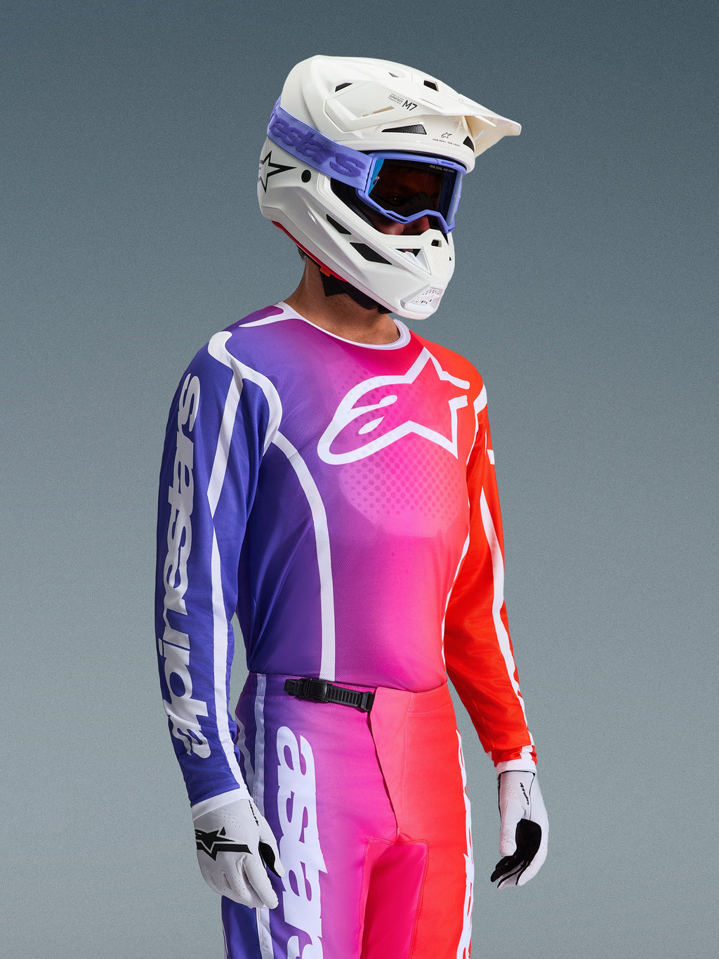Alpinestars 2026 Fluid Apex Jersey, jersey de motocross, degradado vibrante en púrpura, rosa y naranja, usado con pantalones a juego, casco blanco con gafas azules y guantes blancos, diseño atlético para rendimiento y movilidad off-road