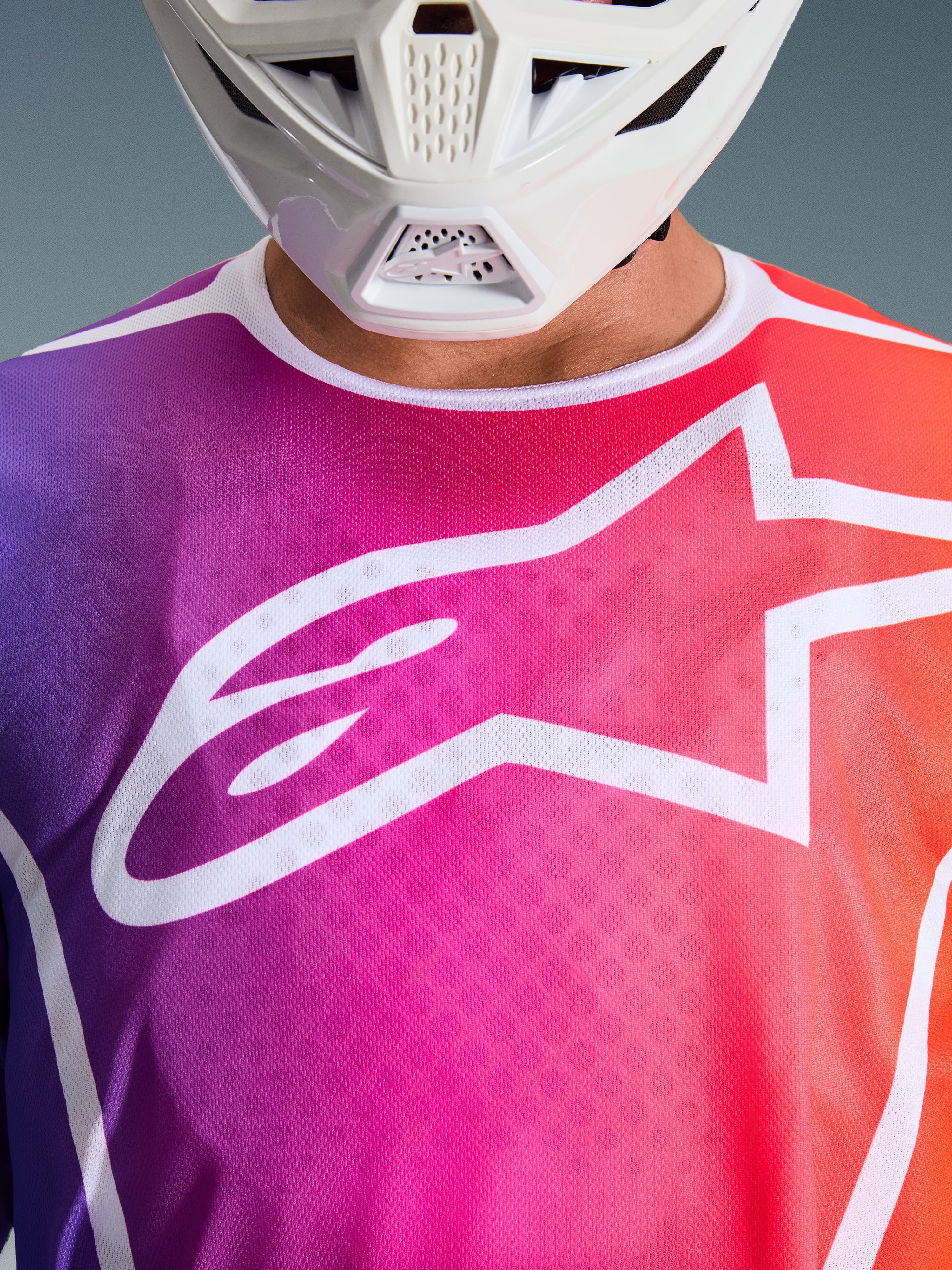 Alpinestars Jersey Fluid Apex 2026, jersey de motocross, degradado multicolor en púrpura, rosa y naranja, con un gran logo de Astars en blanco en el pecho, primer plano usado con un casco de motocross blanco