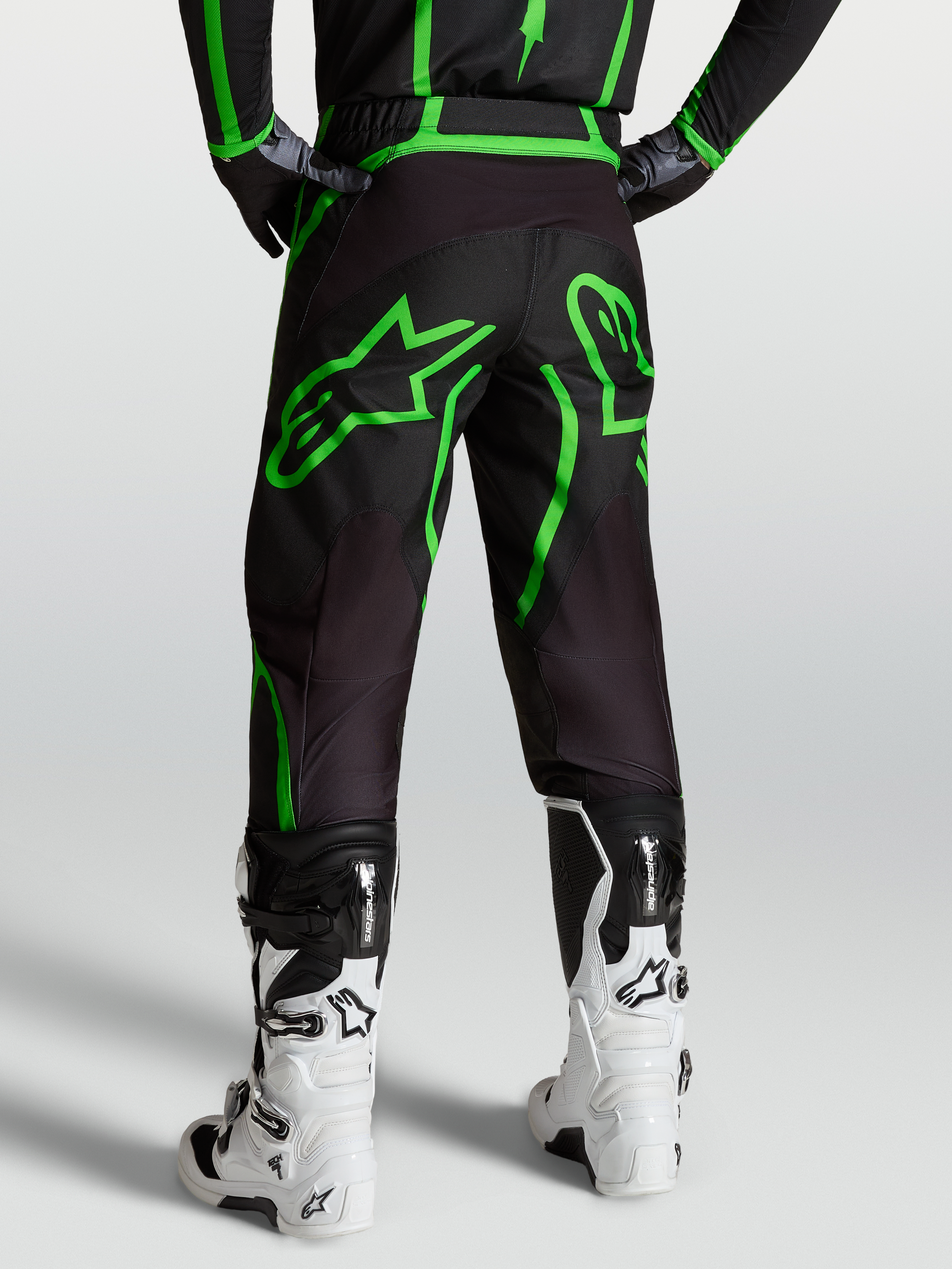 Alpinestars Fluid Apex V2, pantalones de motocross para hombre, negros y verde fluor, vista trasera sobre modelo con botas blancas