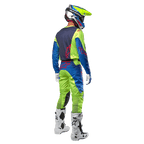 Piloto de motocross visto desde atrás con el jersey Alpinestars Racer Hoen 2024 en amarillo flúor y azul marino, pantalones de motocross a juego y botas blancas, mostrando el diseño posterior con logotipos rojos, equipamiento coordinado para conducción off-road profesional