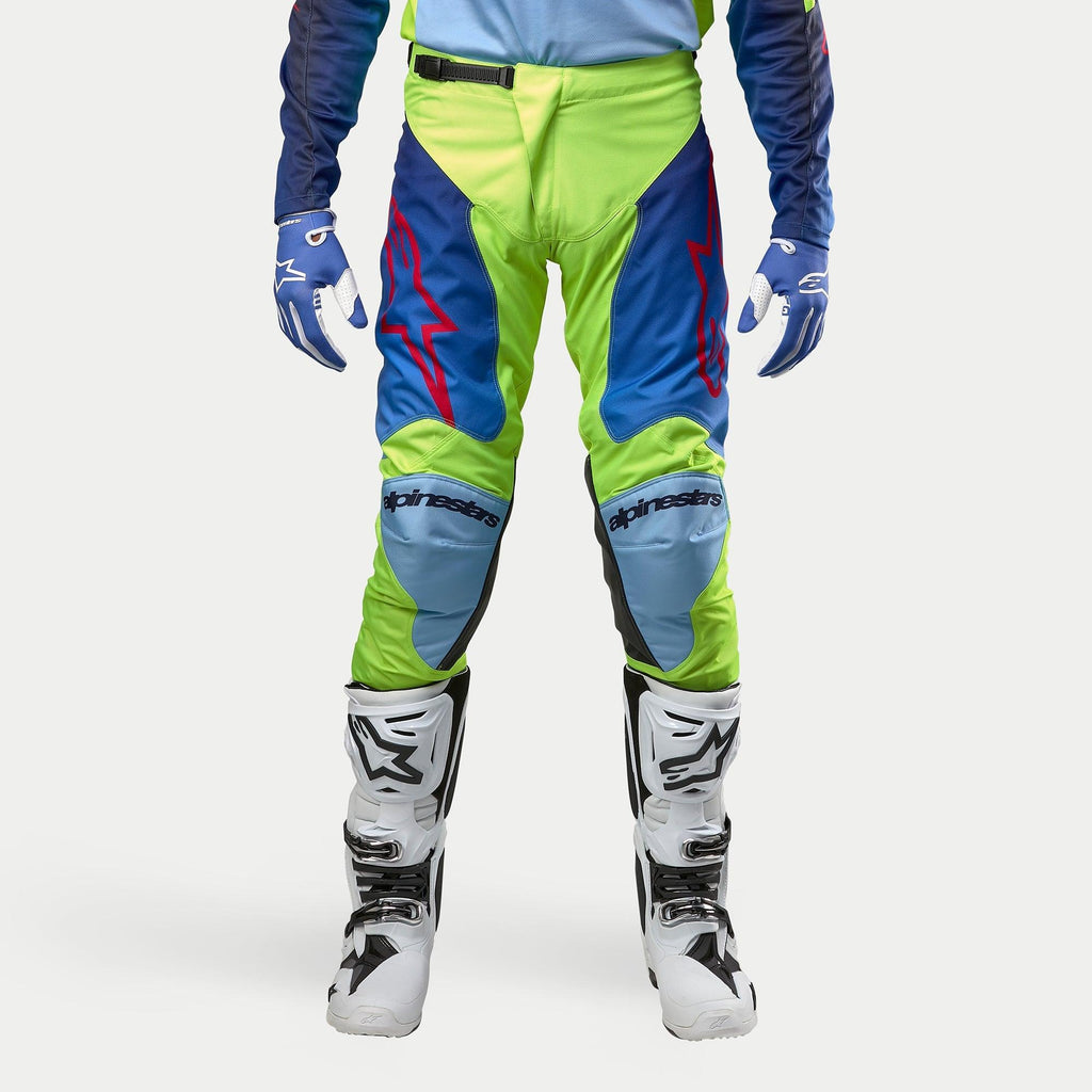 Pantalones Alpinestars Racer Hoen 2024, pantalones de motocross, color amarillo flúor y azul noche, combinados con botas de motocross blancas y negras y guantes azules, con logotipos rojos llamativos, paneles de rodilla en azul claro y un diseño ergonómico deportivo para conducción fuera de carretera