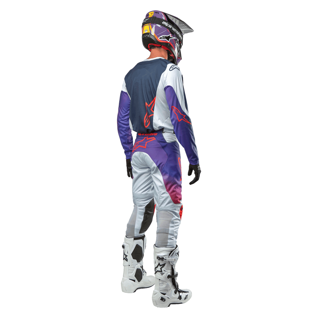 Piloto de motocross visto desde atrás con el jersey Alpinestars Racer Hoen 2024 en Gris Claro Naranja Intenso Negro, panel trasero azul marino con logo de estrella naranja, mangas moradas, combinado con pantalones de carreras a juego y botas blancas
