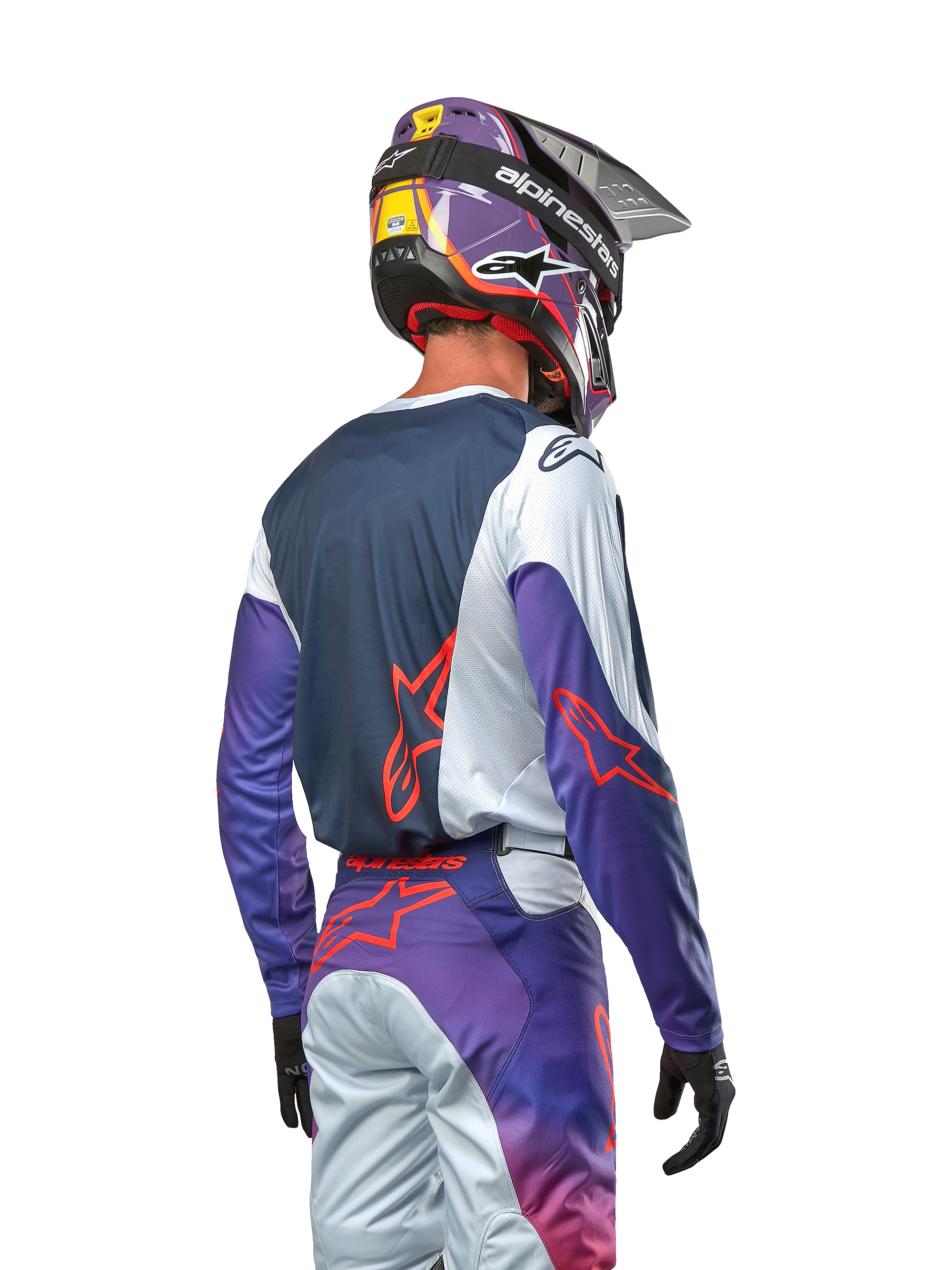 Piloto de motocross visto desde atrás con el Jersey Racer Hoen 2024, panel trasero azul marino con hombros gris claro y mangas moradas, logotipos Alpinestars en naranja intenso, pantalones a juego en morado y gris con casco de motocross morado, equipamiento completo de carreras off-road
