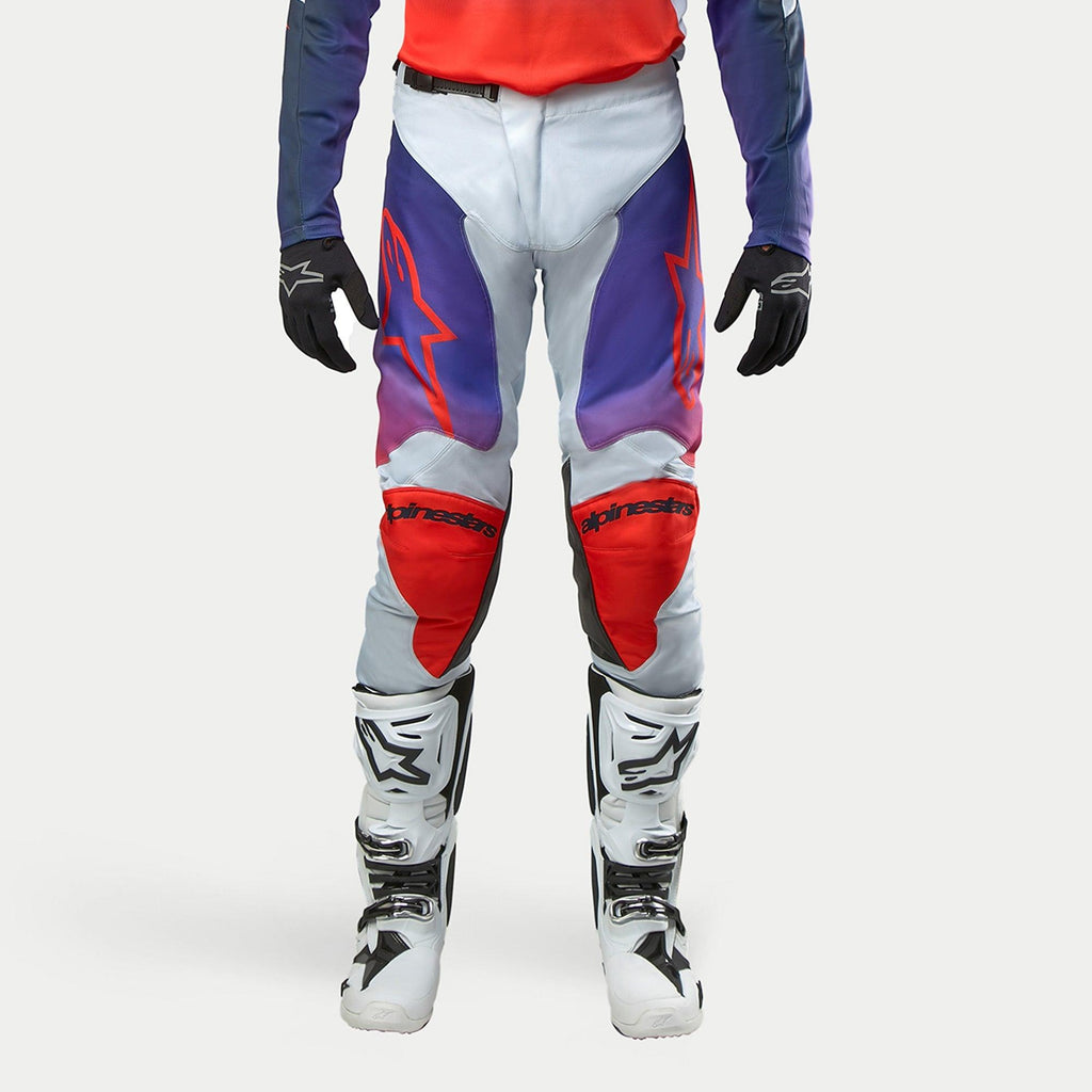 Alpinestars 2024 Racer Hoen Pants, pantalones de motocross para hombre, gris claro con paneles en naranja vibrante, negro y degradados púrpuras, rodilleras rojas con logo, mostrados con botas blancas y guantes negros, diseño técnico para todoterreno.