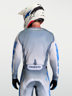 Alpinestars Jersey Racer Amber, jersey de motocross gris y blanco para hombre, vista trasera en modelo con casco blanco, materiales técnicos transpirables y gráficos en azul