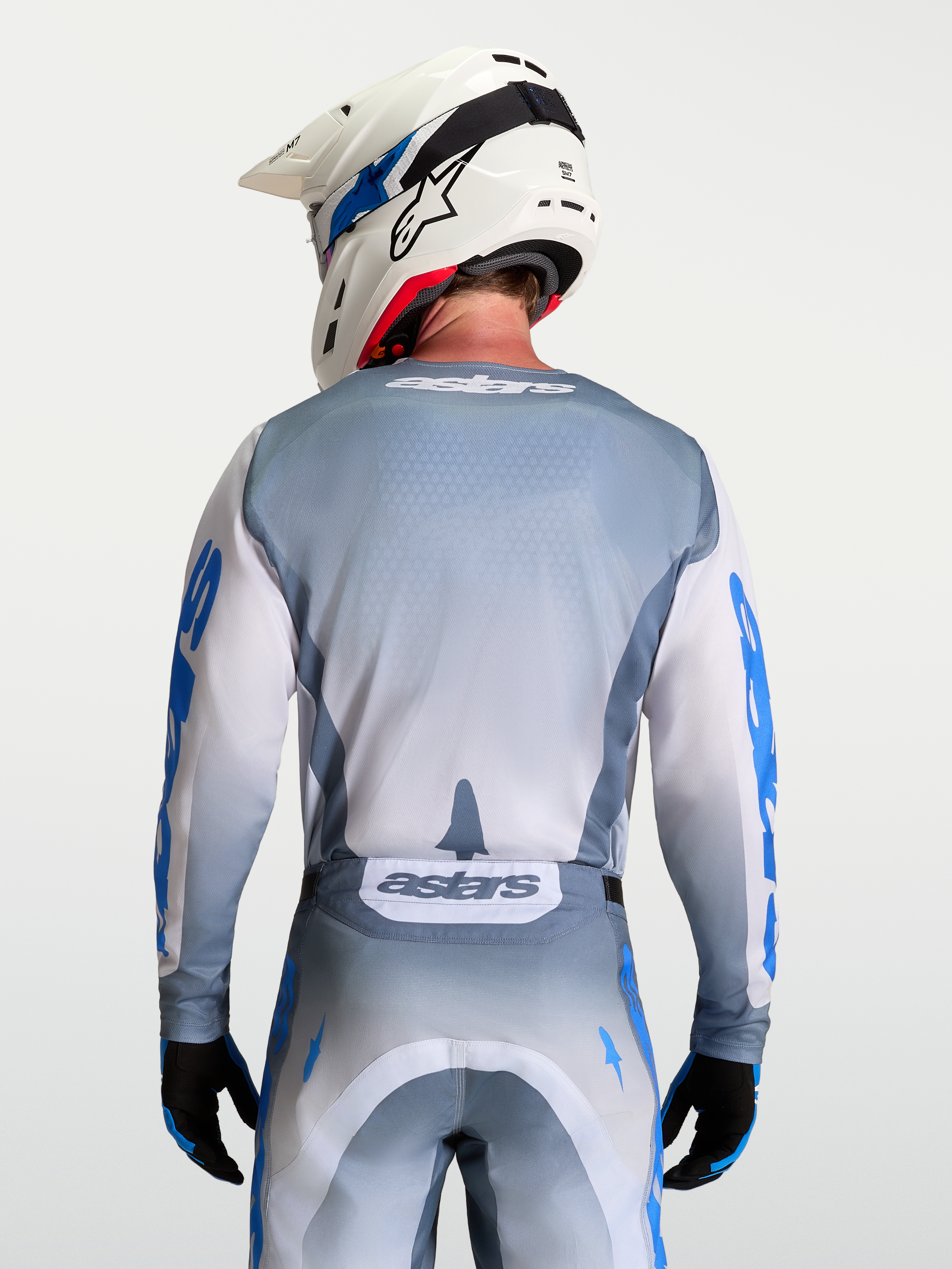 Alpinestars Jersey Racer Amber, jersey de motocross gris y blanco para hombre, vista trasera en modelo con casco blanco, materiales técnicos transpirables y gráficos en azul