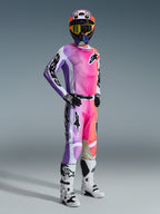 Piloto de motocross con equipamiento de competición completo que incluye los pantalones Alpinestars 2026 Racer Air Portl, diseño multicolor vibrante en rosa, púrpura y naranja con detalles blancos, jersey de manga larga a juego, casco negro con gafas amarillas y botas blancas, conjunto coordinado para carreras off-road de alto rendimiento.