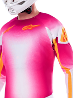 Alpinestars Supertech AFD MIG Edición Limitada, jersey de motocross en degradado rosa flúor y blanco, con logotipos Astars y detalles en las mangas en color naranja, usado por un piloto con casco blanco, ajuste atlético de competición