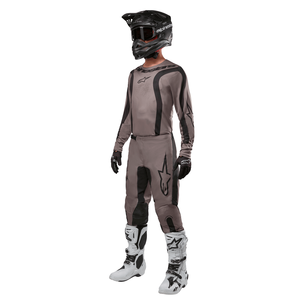 Piloto de motocross con equipamiento completo de carreras que incluye el jersey Alpinestars Fluid Lurv 2024 en color Mud Black, pantalones a juego, casco negro con gafas, botas blancas y negras, y guantes negros, conjunto coordinado para carreras todoterreno