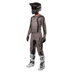 Piloto de motocross con equipamiento completo de carreras que incluye el jersey Alpinestars Fluid Lurv 2024 en color Mud Black, pantalones a juego, casco negro con gafas, botas blancas y negras, y guantes negros, conjunto coordinado para carreras todoterreno