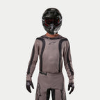 Piloto de motocross con el Jersey Alpinestars 2024 Fluid Lurv en color Mud Black, un tejido gris terroso con franjas gráficas negras y un logo de estrella en el pecho, combinado con casco negro, gafas rojas y guantes negros para un look de carreras todoterreno coordinado.