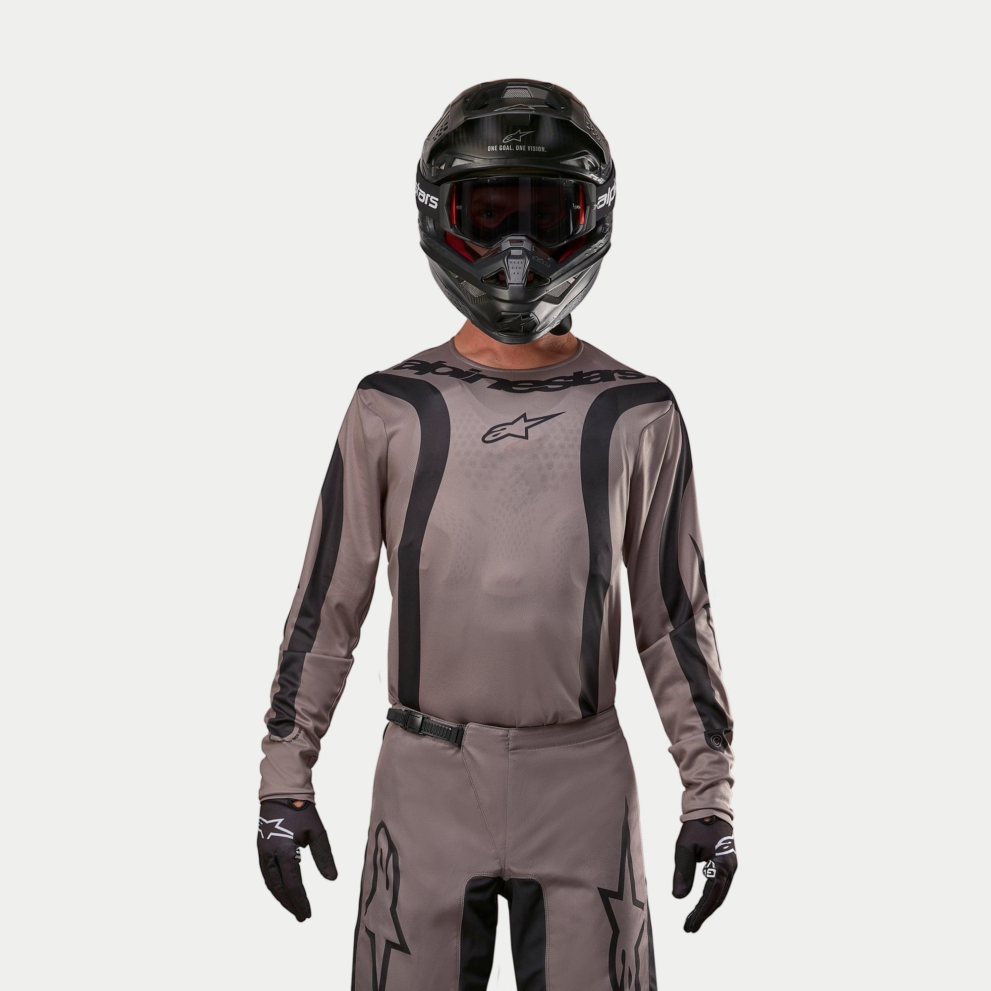 Piloto de motocross con el Jersey Alpinestars 2024 Fluid Lurv en color Mud Black, un tejido gris terroso con franjas gráficas negras y un logo de estrella en el pecho, combinado con casco negro, gafas rojas y guantes negros para un look de carreras todoterreno coordinado.
