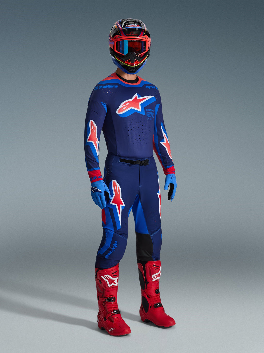 Motocross rider wearing complete racing gear, 2026 Supertech Vista Jersey in Dark Navy with Hot Coral and Cobalt accents, matching motocross pants, multi-colored helmet with red goggles, bright red boots, blue gloves, coordinated outfit for off-road racing