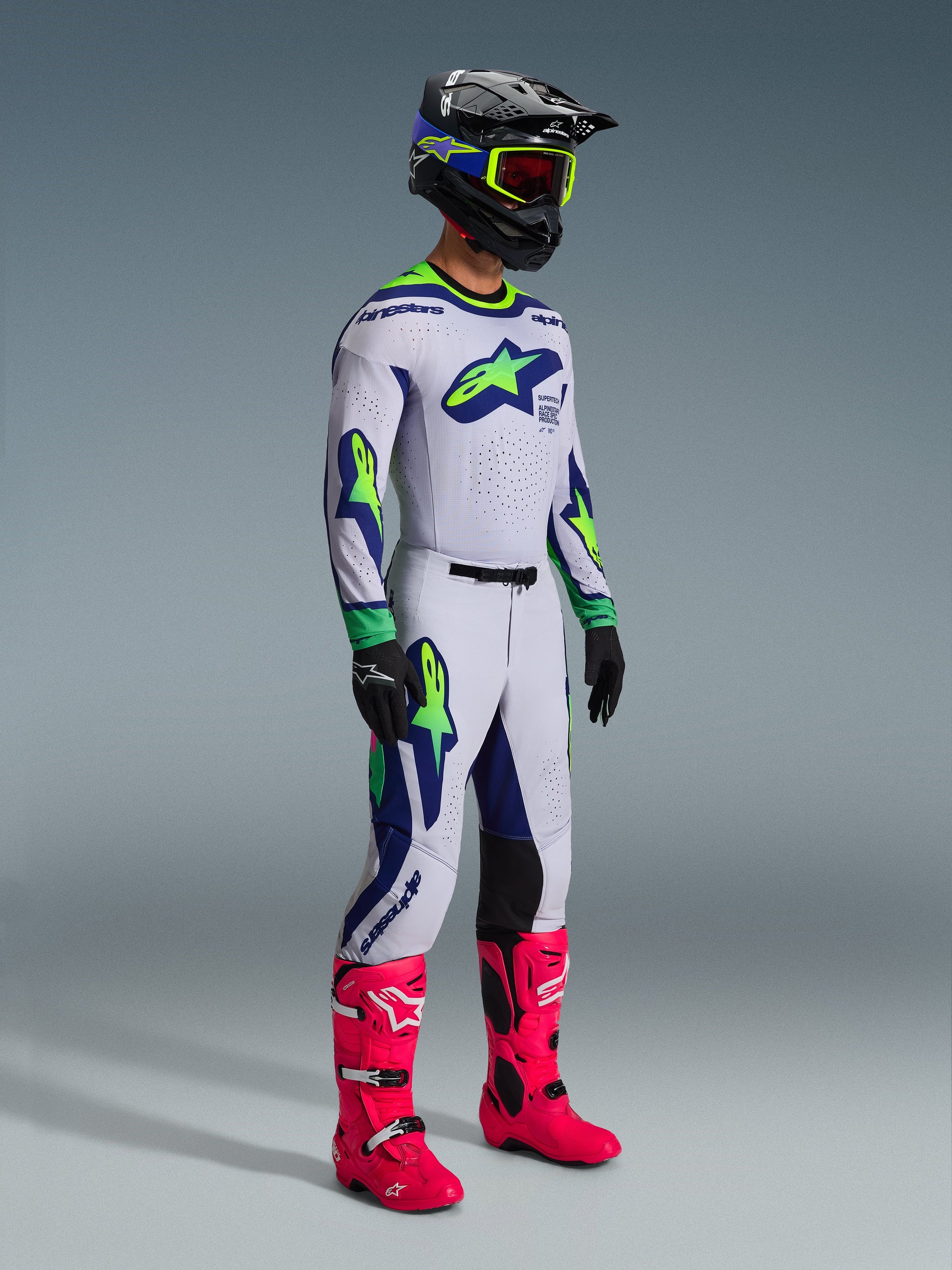 Motocross rider wearing the Alpinestars 2026 Supertech Vista Jersey in Light Gray Yellow Fluo Blue, matching racing pants, bright pink boots, and a black helmet with yellow goggles, featuring laser-cut ventilation and a professional athletic fit for off-road racing