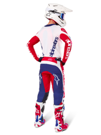 Piloto de motocross visto desde atrás vistiendo el Jersey Alpinestars Techstar Fortitude Edición Limitada, en color rojo brillante, blanco y azul, pantalones técnicos a juego con logotipos de estrellas, casco blanco con gafas, y botas rojas y blancas, equipamiento completo coordinado para carreras off-road