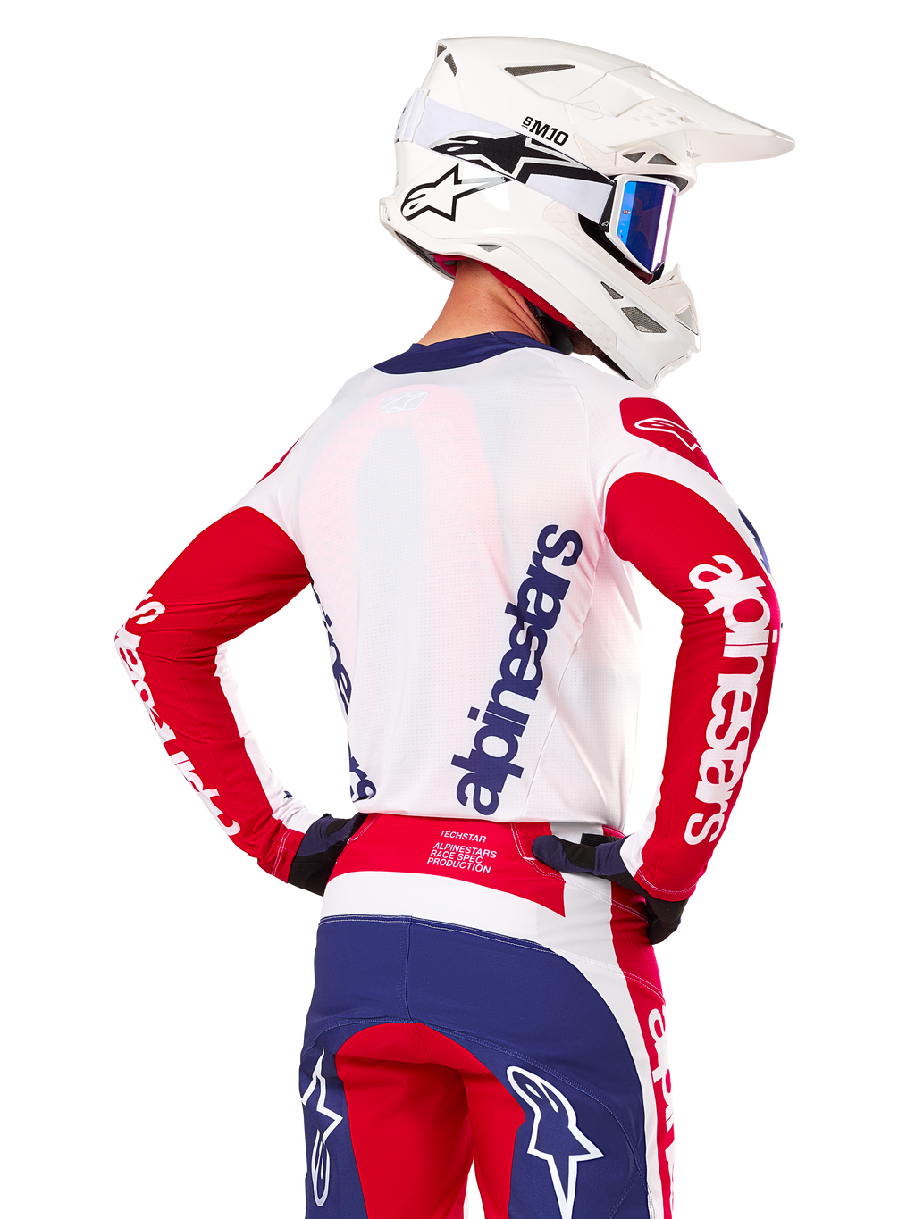 Jersey Alpinestars Techstar Fortitude Edición Limitada, jersey de motocross, rojo brillante, blanco y azul, vista trasera de un piloto con casco blanco y equipamiento a juego, con logotipos llamativos de Alpinestars en la espalda y las mangas, diseñado para carreras off-road de alto rendimiento.